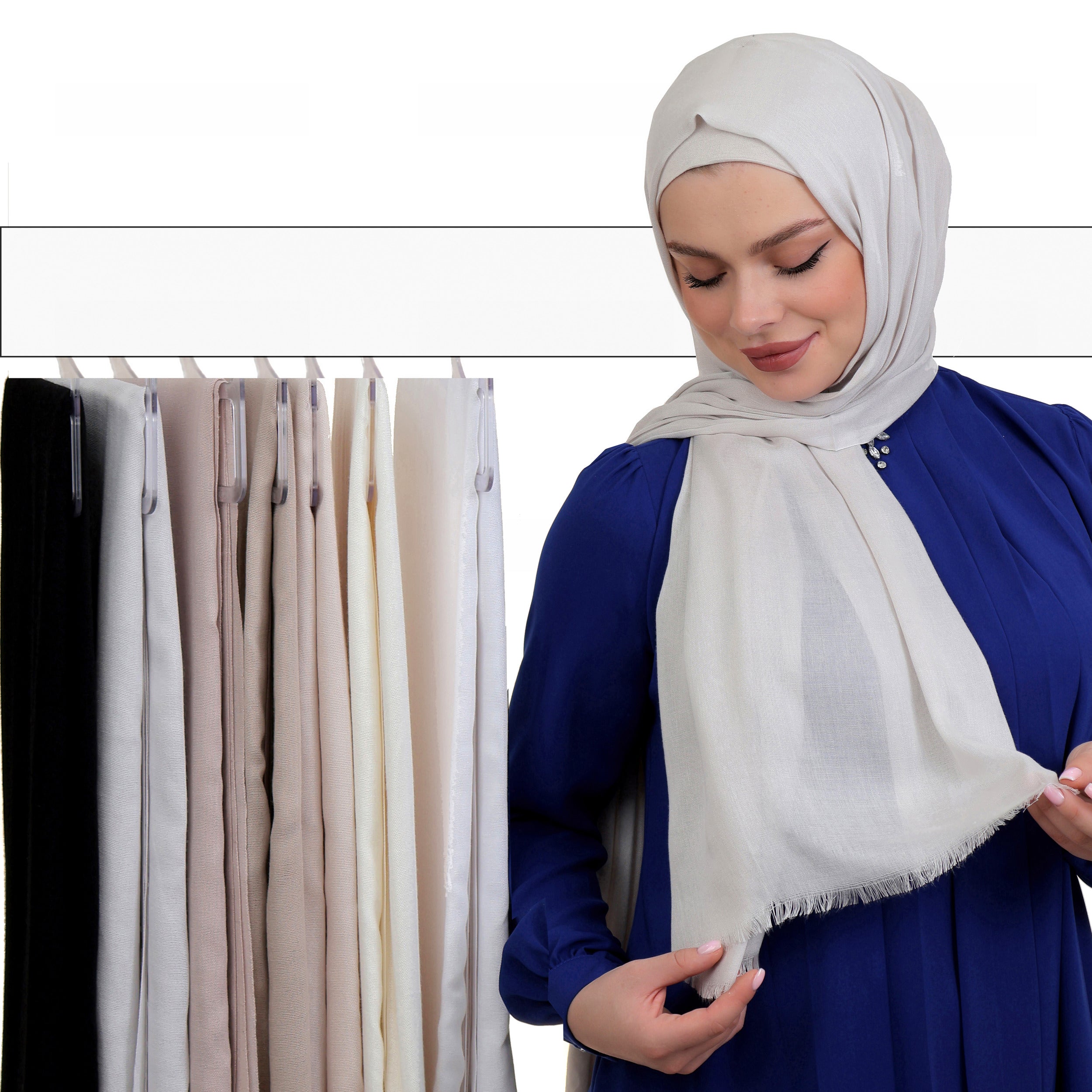 Model wearing an off-white hijab with a royal blue top, standing near a display of folded hijabs in neutral shades.