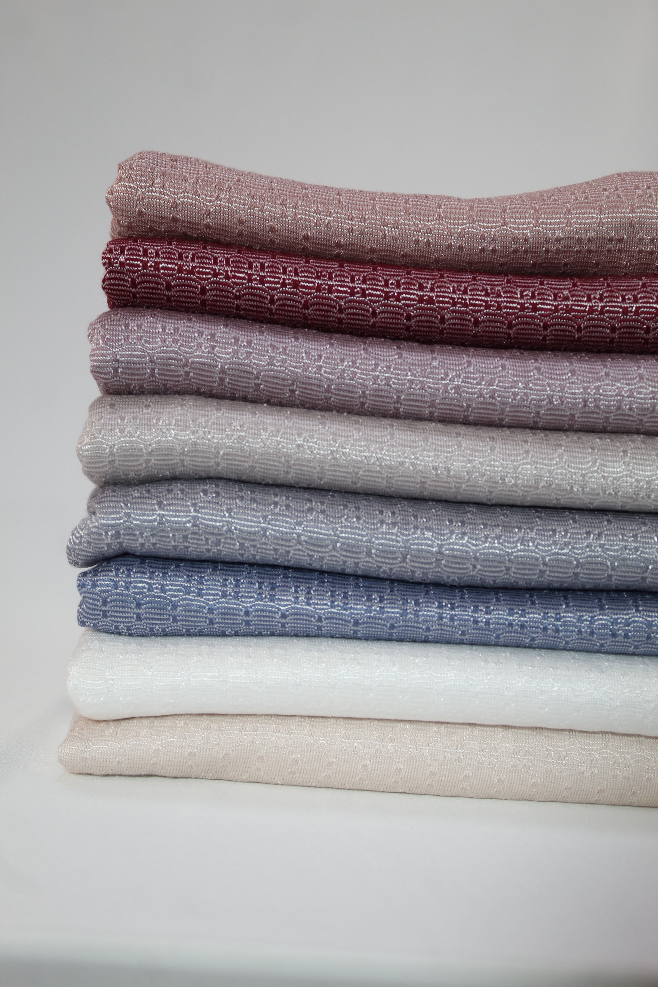 Stacked hijabs in rich earthy colors, including maroon, grey, and cream, highlighting their woven texture.