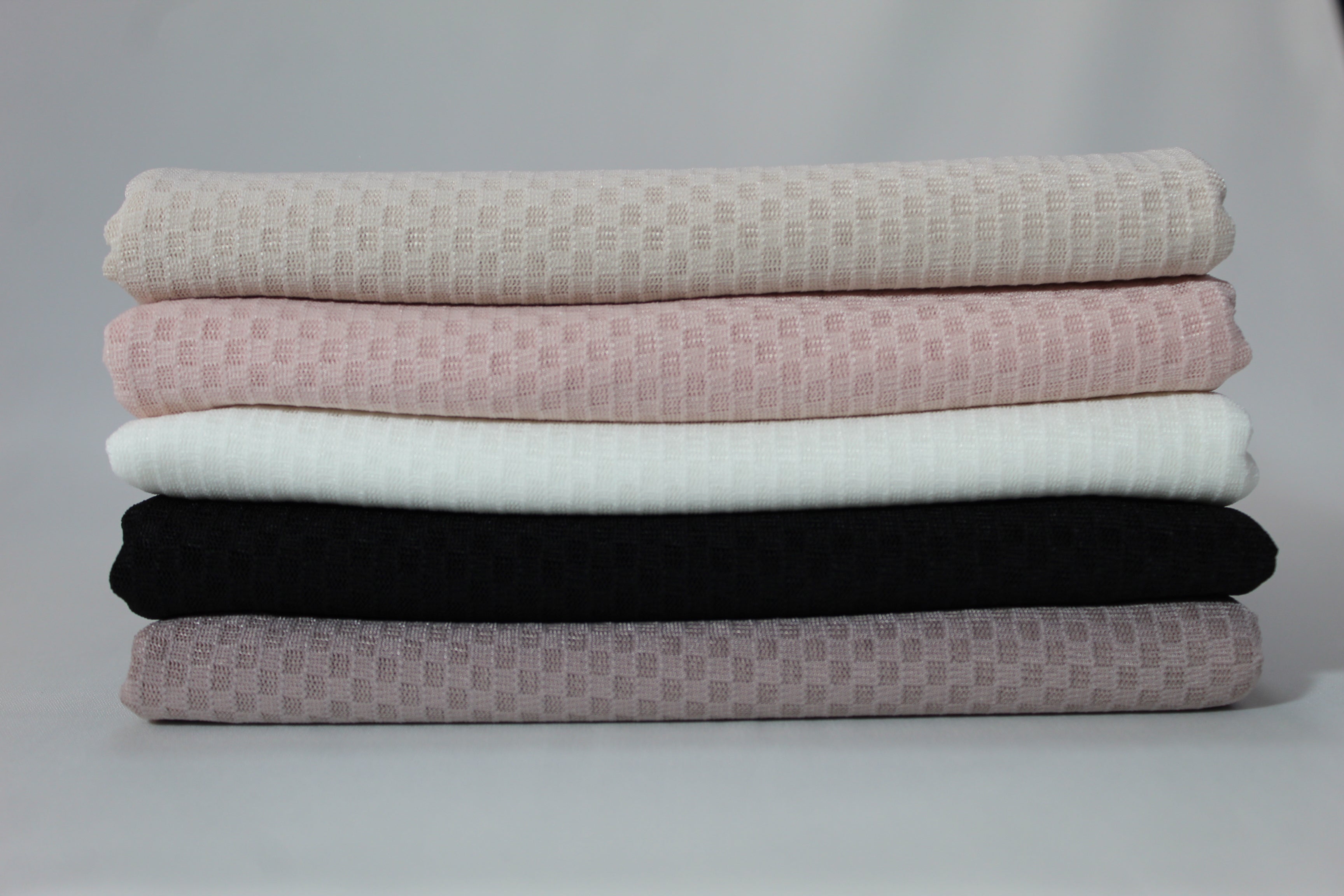Stack of folded hijabs in neutral tones, featuring beige, blush pink, white, black, and taupe colors, showcasing soft textured fabric against a minimalist grey background