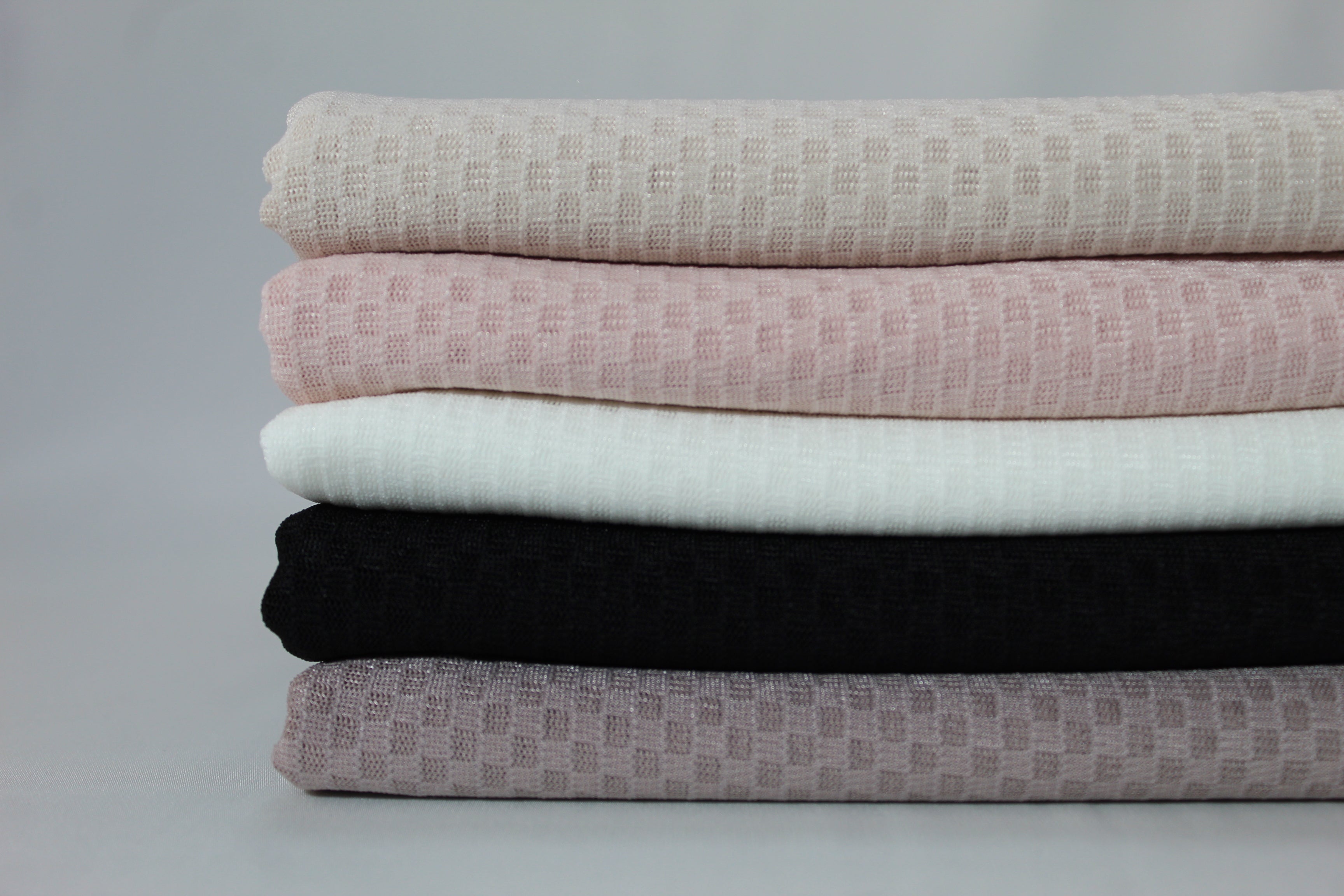Close up picture of a Stack of folded hijabs in neutral tones, featuring beige, blush pink, white, black, and taupe colors, showcasing soft textured fabric against a minimalist grey background