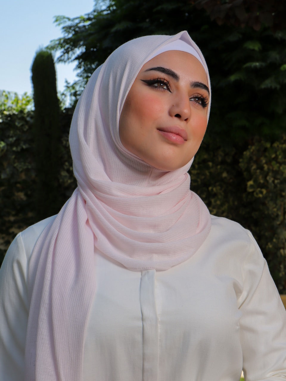 Model in a pale pink hijab posing outdoors, exuding elegance and modesty.