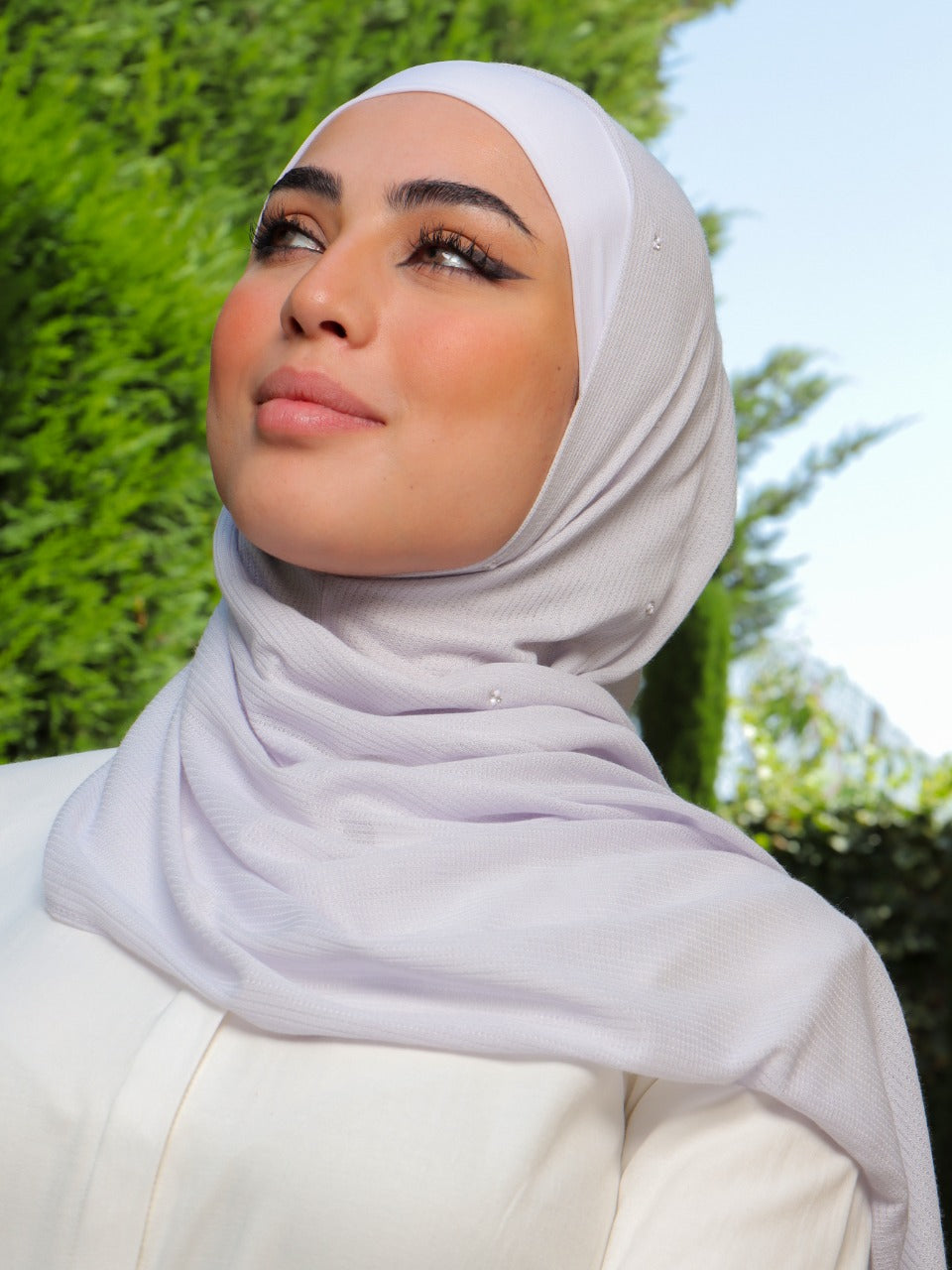 Model in a lavender hijab smiling under the sunlight in a serene outdoor setting.