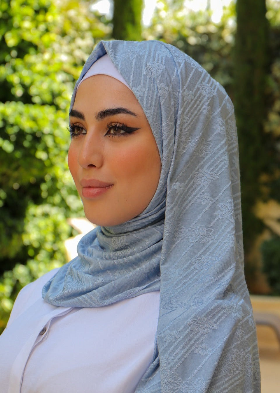 Side view of a woman in a light blue hijab with flower print standing outdoors, with greenery in the background.